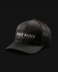 A black trucker-style baseball cap with a structured front panel, black mesh back, and a rectangular front patch reading “HANDS RAISED” with smaller text “WORK. GRIT.