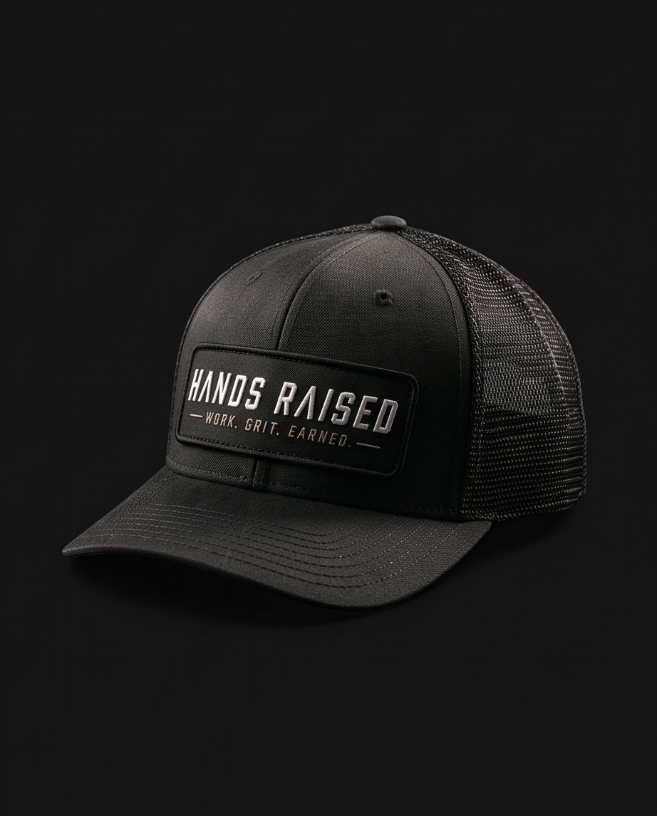 A black trucker-style baseball cap with a structured front panel, black mesh back, and a rectangular front patch reading “HANDS RAISED” with smaller text “WORK. GRIT.