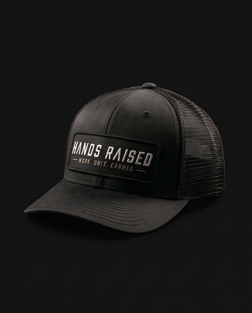 A black trucker-style baseball cap with a structured front panel, black mesh back, and a rectangular front patch reading “HANDS RAISED” with smaller text “WORK. GRIT.
